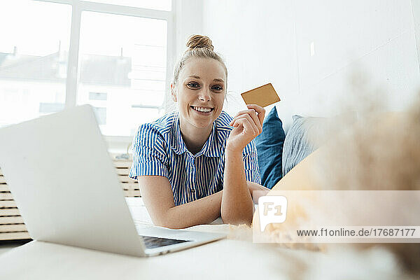 Lächelnder Freiberufler zeigt Kreditkarte per Laptop  der zu Hause im Bett liegt