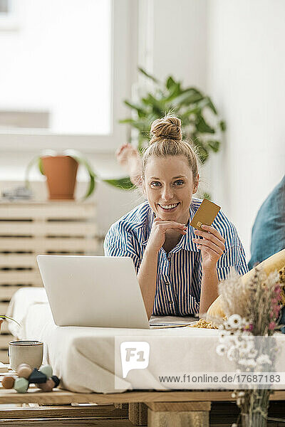 Businesswoman holding credit card by laptop lying on bed at home