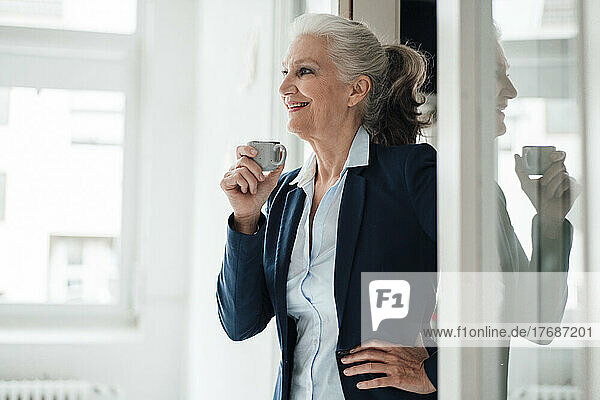 Glückliche Geschäftsfrau hält Kaffeetasse und steht mit der Hand auf der Hüfte im Büro