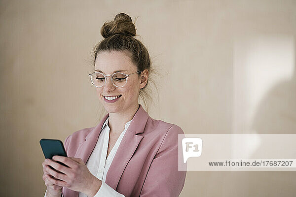 Businesswoman wearing eyeglasses using smart phone by wall