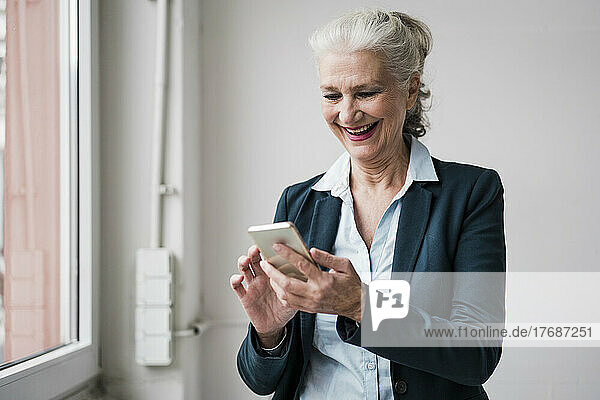 Happy businesswoman using smart phone standing by window in office