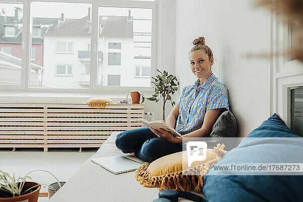 Smiling young freelancer with book sitting on bed at home