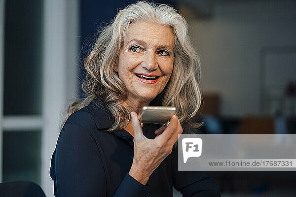 Happy senior woman sending voicemail through smart phone in office