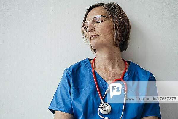 Exhausted female doctor with eyes closed leaning on white wall resting in hospital