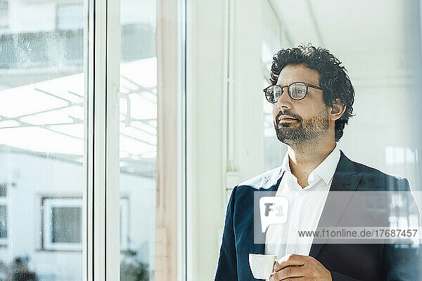 Thoughtful businessman holding coffee cup looking through window in office