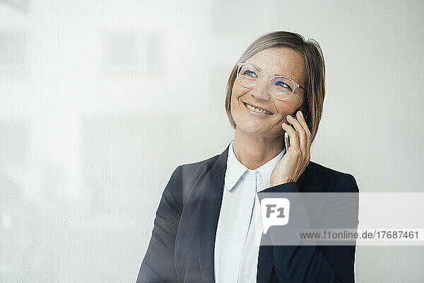 Happy businesswoman wearing blazer talking on smart phone seen through glass window