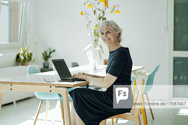 Happy businesswoman with laptop sitting on chair at desk in office