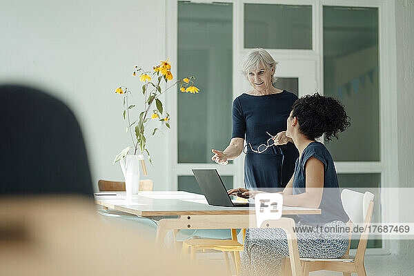 Senior businesswoman discussing with colleague sitting at desk in office