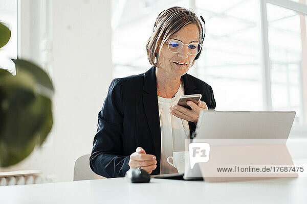 Smiling telecaller with smart phone using tablet PC sitting at desk in office