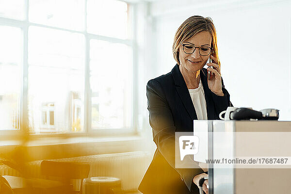 Businesswoman talking on smart phone using coffee maker in office cafeteria