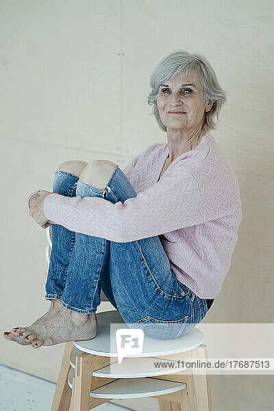 Smiling senior woman with gray hair sitting on chairs in front of wall