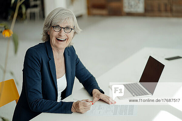 Cheerful businesswoman with document sitting at desk in office