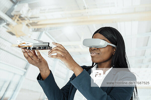 Lächelnde Geschäftsfrau trägt einen Virtual-Reality-Simulator und hält Maschinenteile in der Fabrik
