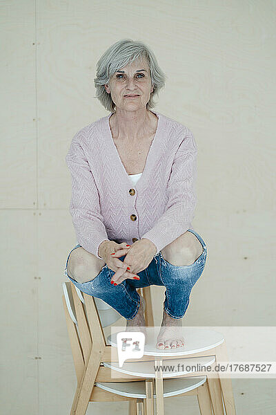 Smiling senior woman with gray hair crouching on chairs in front of wall