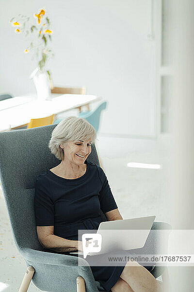 Lächelnde ältere Frau mit Laptop im Sessel im Büro sitzend