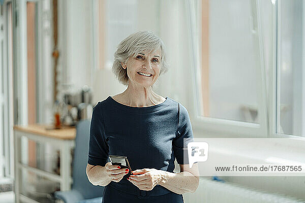 Smiling senior businesswoman with mobile phone standing in office