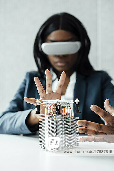 Businesswoman wearing virtual reality simulator gesturing at machine part on desk