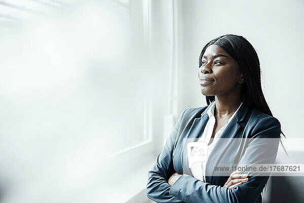 Young businesswoman looking through window standing with arms crossed in office