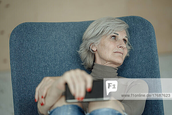 Nachdenkliche ältere Frau mit Smartphone sitzt auf einem Stuhl vor der Wand