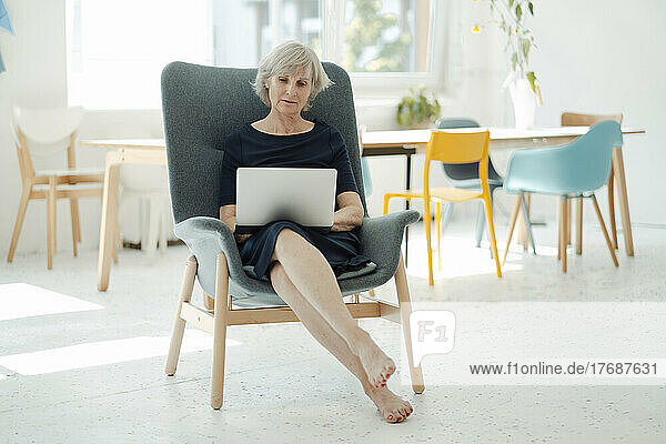 Senior businesswoman using laptop sitting in armchair in office
