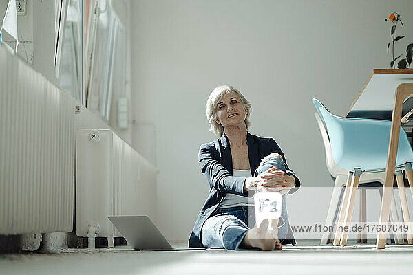 Smiling senior businesswoman with laptop sitting on ground in office
