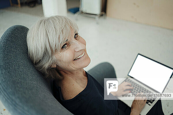 Happy senior businesswoman with laptop sitting in armchair in office