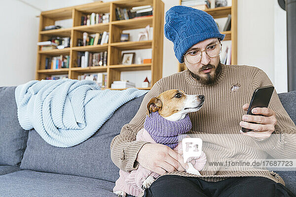Man with wooly hat sitting on couch holding dog checking smartphone