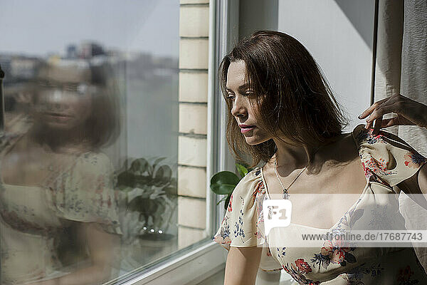 Mature woman with brown hair standing by glass window at home