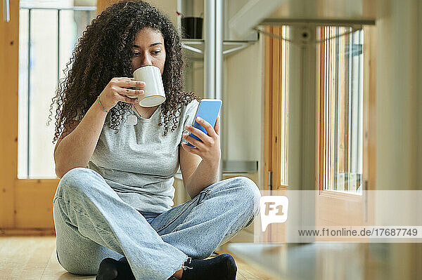 Young woman drinking coffee and using mobile phone sitting on floor at home