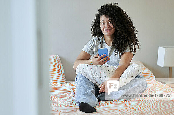 Smiling woman holding mobile phone sitting on bed at home