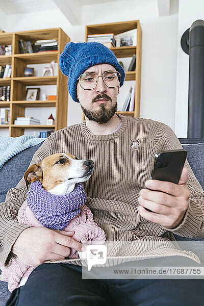 Man with wooly hat sitting on couch holding dog checking smartphone