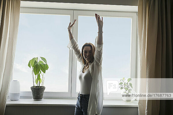 Happy mature woman with arms raised standing in front of window