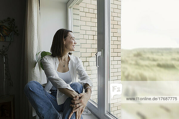 Smiling mature woman sitting on window sill at home