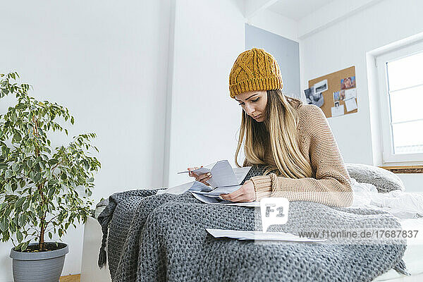 Woman with wooly hat sitting on bed checking bills