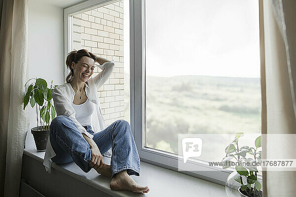 Happy mature woman with hand in hair sitting on window sill at home