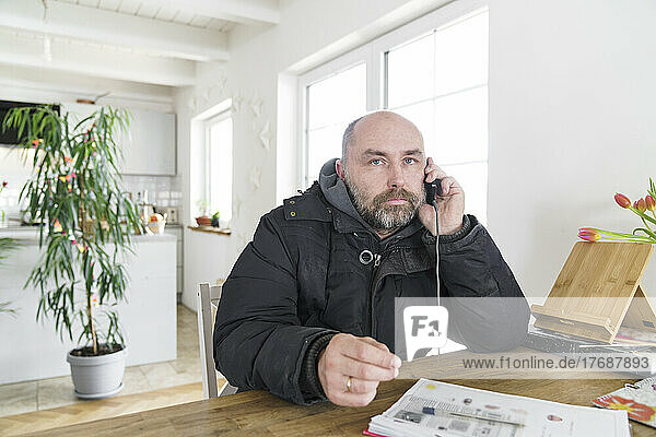Reifer Mann sitzt zu Hause in Winterjacke und telefoniert
