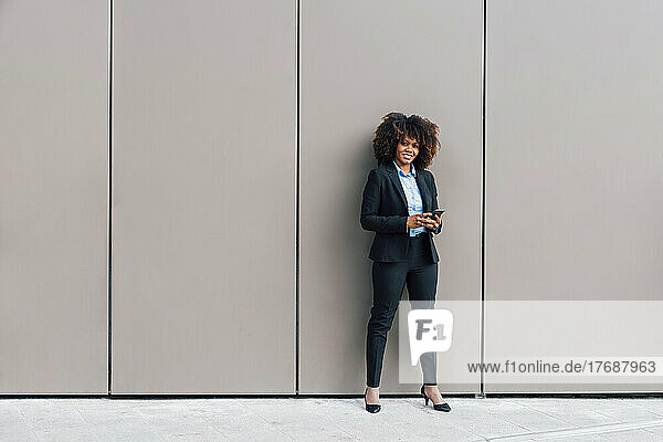 Smiling businesswoman holding mobile phone standing in front of wall