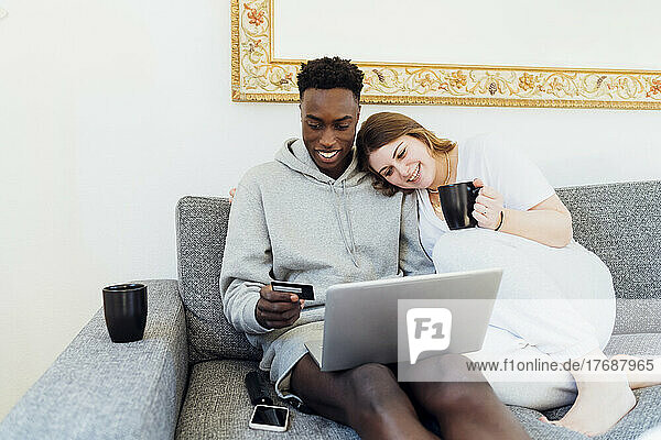 Happy young couple with credit card and laptop sitting together on sofa at home