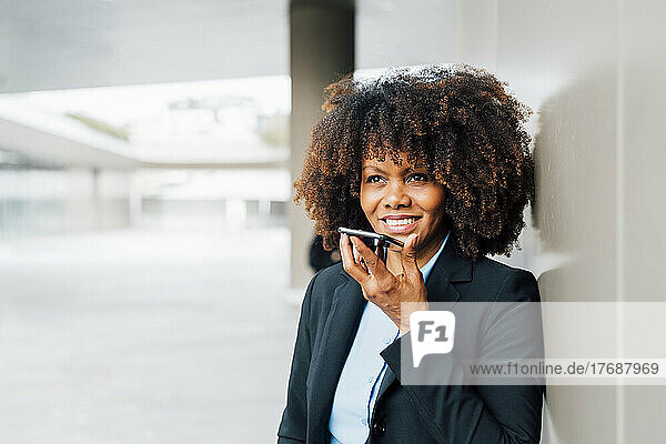 Lächelnde Geschäftsfrau spricht über den Smartphone-Lautsprecher  der an der Wand lehnt