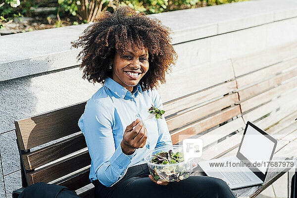 Glückliche Geschäftsfrau isst Salat und sitzt mit Laptop auf der Bank