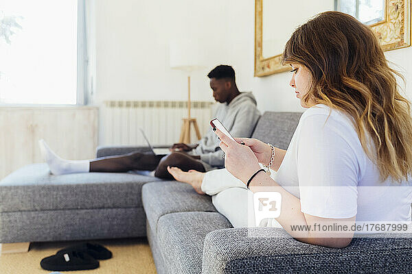 Junge Frau benutzt Smartphone und sitzt mit ihrem Freund zu Hause auf dem Sofa