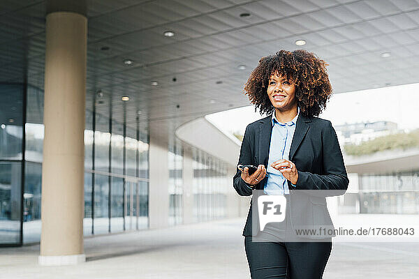 Smiling businesswoman holding smart phone walking outside office building
