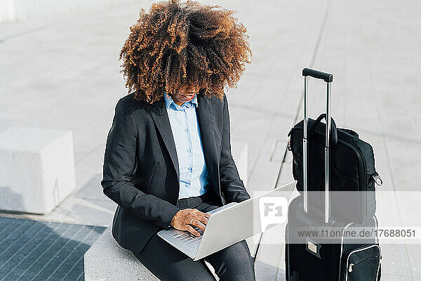 Afro Businesswoman using laptop sitting on concrete block by luggage
