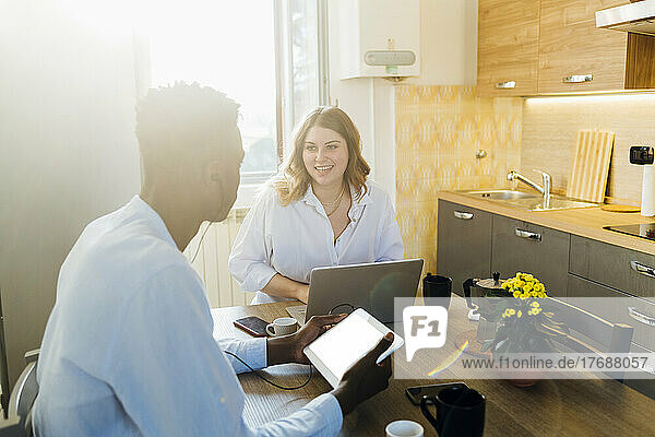 Smiling young woman with laptop sitting by boyfriend at dining table in kitchen