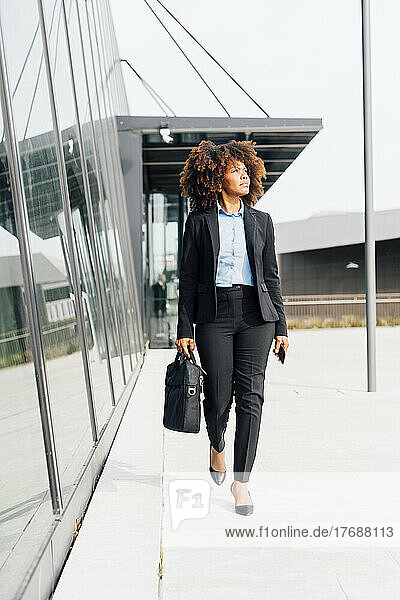 Businesswoman holding laptop bag walking by glass wall