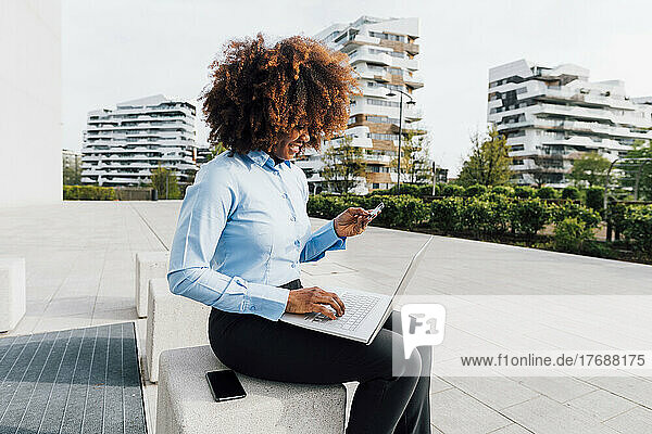 Lächelnde Geschäftsfrau mit Kreditkarte beim Online-Einkauf per Laptop auf Betonblock sitzend