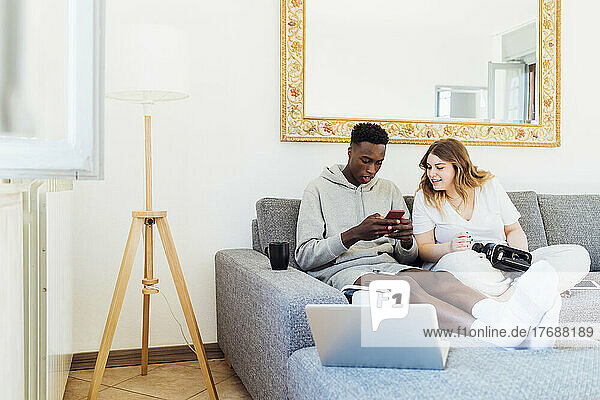 Young man using smart phone sitting by girlfriend in living room at home