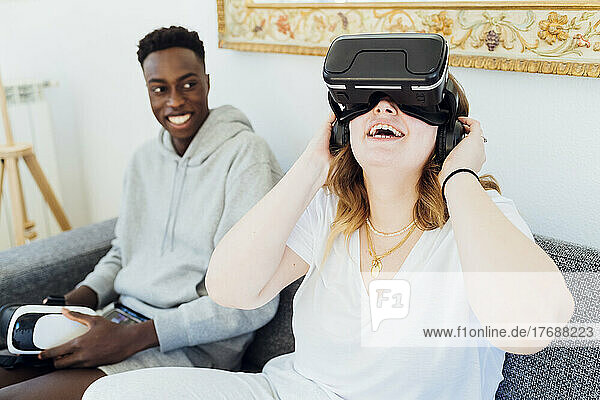 Happy woman wearing virtual reality simulator sitting with boyfriend on sofa at home