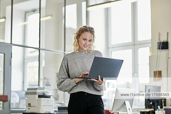 Smiling young woman standing in office using laptop