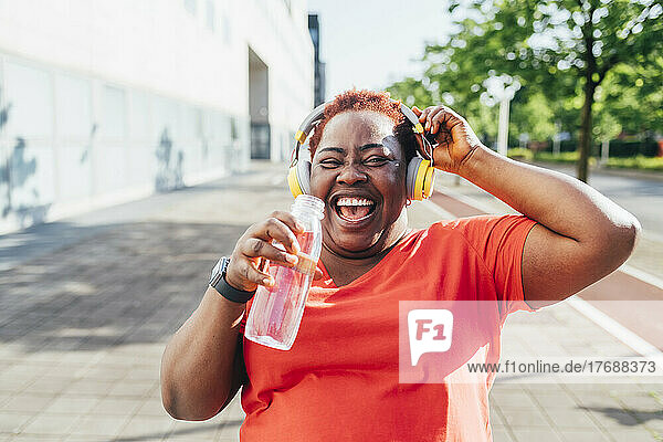 Cheerful woman holding water bottle listening music through wireless headphones